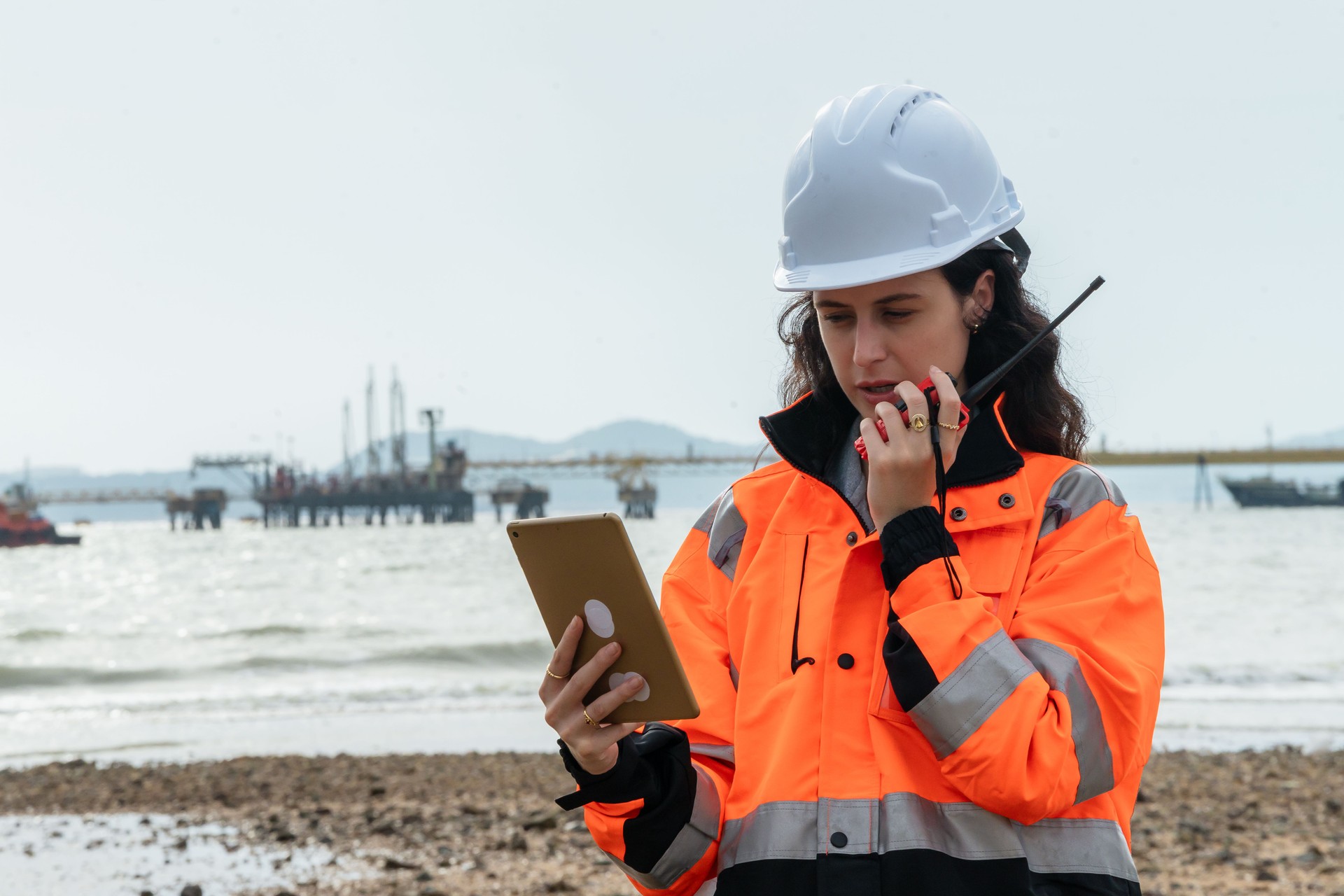 Female engineer using a tablet and walkie-talkie for a field report at a maritime construction site. Woman technician multitasking with mobile technology for data entry and communication offshore.